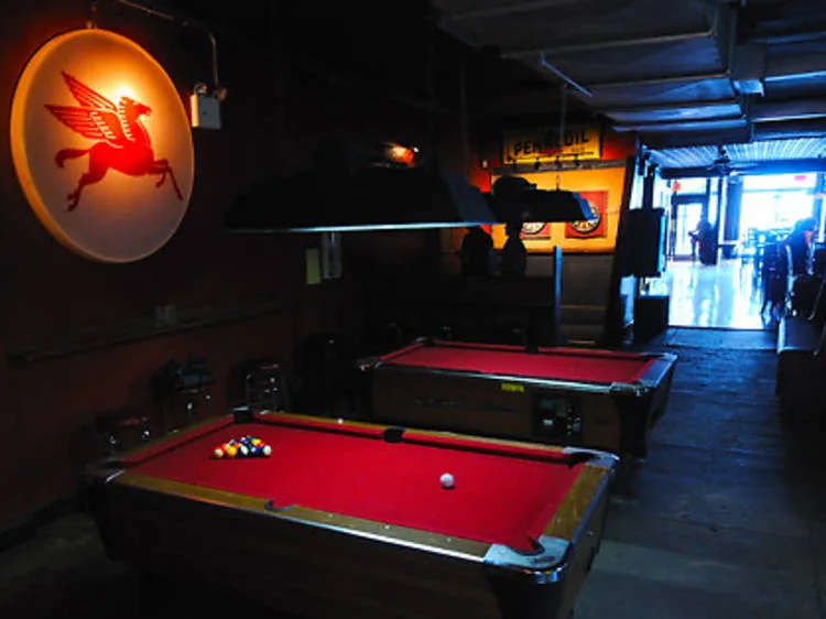 Two red-felt pool tables under low hanging lamps at Ace Bar, with a glowing red flying-horse sign on the brick wall and the long room stretching toward the entrance.